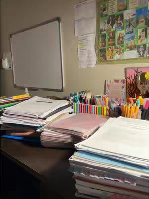 A desk is covered in a messy arrangement of school supplies and papers. Piles of documents, notebooks, and binders are scattered across the surface, along with colorful pens, markers, and highlighters. A white board is visible on the wall. The overall composition conveys a sense of organized chaos, indicative of a study session or academic work.