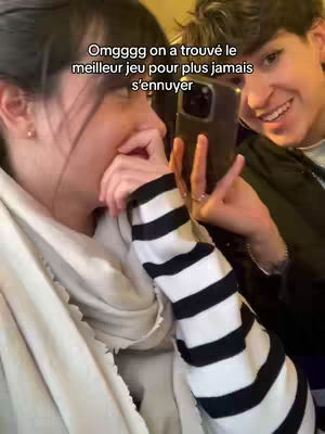 Two people are taking a selfie with a smartphone. The woman on the left is partially in the foreground, covering her mouth with her hand, and wearing a black and white striped sleeve and a light-colored scarf. The other person to the right is smiling. The background is slightly blurred, suggesting an indoor or low-light setting, and the overlaid text "Omgggg on a trouvé le meilleur jeu pour plus jamais s'ennuyer" appears at the top of the image.