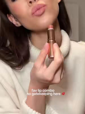 A close-up shot featuring a woman holding a lipstick tube near her lips. The image is well-lit, with a soft focus on the woman's face and the lipstick. The background is blurred, helping to highlight the product and the woman's features.