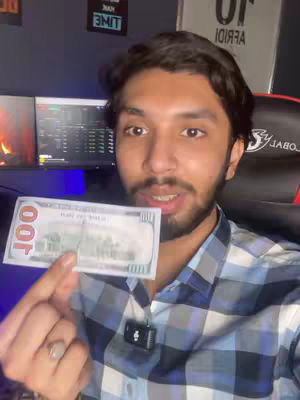 A man with dark hair and a beard is holding up a hundred-dollar bill in front of his face. He is smiling and looking at the camera. Behind him, there are two computer monitors displaying data, as well as a gaming chair and the top of a sign.