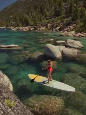 A scenic shot of a woman paddleboarding on clear turquoise water, surrounded by rocks and a forested mountain in the background.