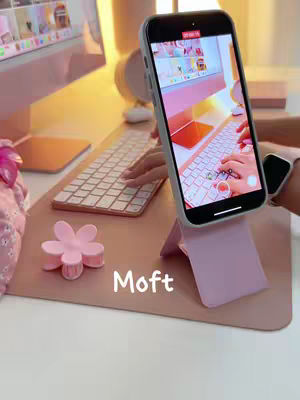 The cover image shows a desk setup with a pink theme. A phone is propped up on a pink stand, displaying a screen recording. A white keyboard, a pink flower-shaped item, and a computer monitor are also visible. The word "Moft" is written in white on the table. A person's arm is visible holding the phone.