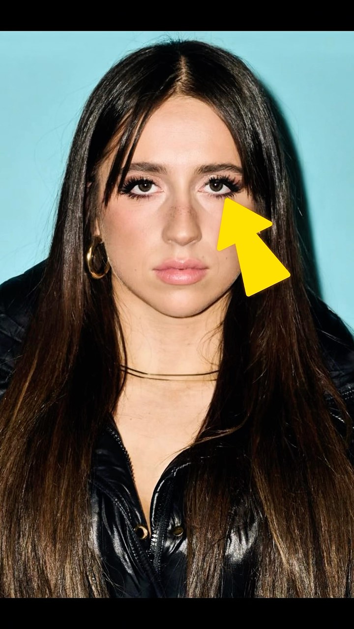 A close-up portrait of a woman with long dark hair and a striking gaze. The background is a solid light blue. The woman is facing the camera directly, and her expression is slightly neutral. She wears a black jacket and gold jewelry. A yellow arrow points at her eye.