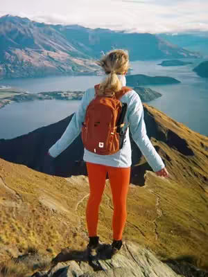 A woman stands on a mountain peak with her back to the camera, arms outstretched, overlooking a scenic landscape of mountains and water. She wears orange pants, a white top, and a backpack.