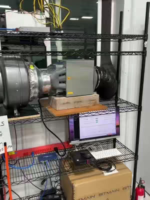 The image shows a home crypto mining setup. A computer screen displays mining software, and cabling and additional hardware is connected to a large fan.