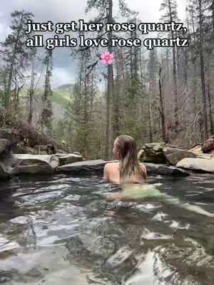 A woman stands in a natural pool of water, facing away from the camera towards a backdrop of a lush forest. The image has cool tones, with the water reflecting the sky and the surrounding trees. Text overlayed on the top saying "just get her rose quartz, all girls love rose quartz". A pink flower is placed in the center of the frame.
