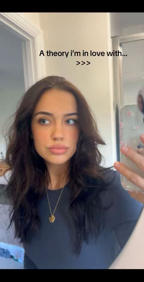 A woman is taking a selfie in a bathroom with a mirror in the background. The woman is looking towards the camera, has long wavy brown hair, and is wearing a dark shirt and a gold necklace. The text overlay says, "A theory I'm in love with >>>"