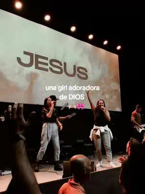 A live performance scene in a large hall. Two female singers are on a brightly lit stage, one holding a microphone and the other playing a guitar. Behind them, a large screen displays the word 'JESUS' in bold letters. A crowd of people is visible, some with raised hands, suggesting a worship or concert atmosphere.
