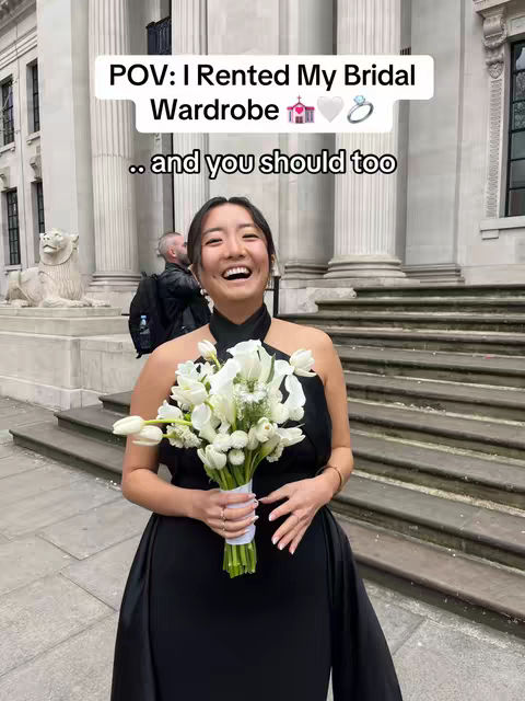 A woman with dark hair smiles, holding a bouquet of white flowers in front of a white background with stairs. Text overlaid says "POV: I Rented My Bridal Wardrobe .. and you should too" in white text within a rounded rectangle over the woman's head.