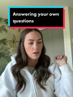 A woman with long brown hair stands in front of a patterned wall and a white door. She wears a white hoodie and looks towards the camera with a neutral expression. A black banner with white text that says 'Answering your own questions' is placed above her head.