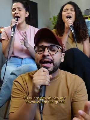The image shows a man in the foreground wearing glasses and a maroon cap, holding a microphone and singing. Behind him, there are two women with microphones singing as well. They are indoors with some background elements.