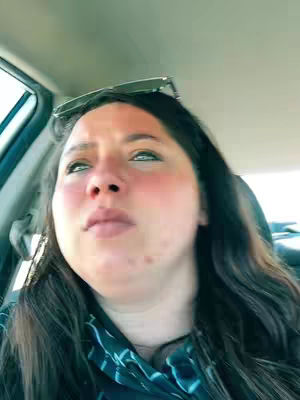 A selfie-style shot from inside a car, featuring a woman looking upwards with a slightly blurred expression. Her dark hair is parted on the side, and sunglasses rest above her forehead. The lighting is soft, highlighting her face with hints of reddish blush, and faint scenery can be glimpsed behind her.