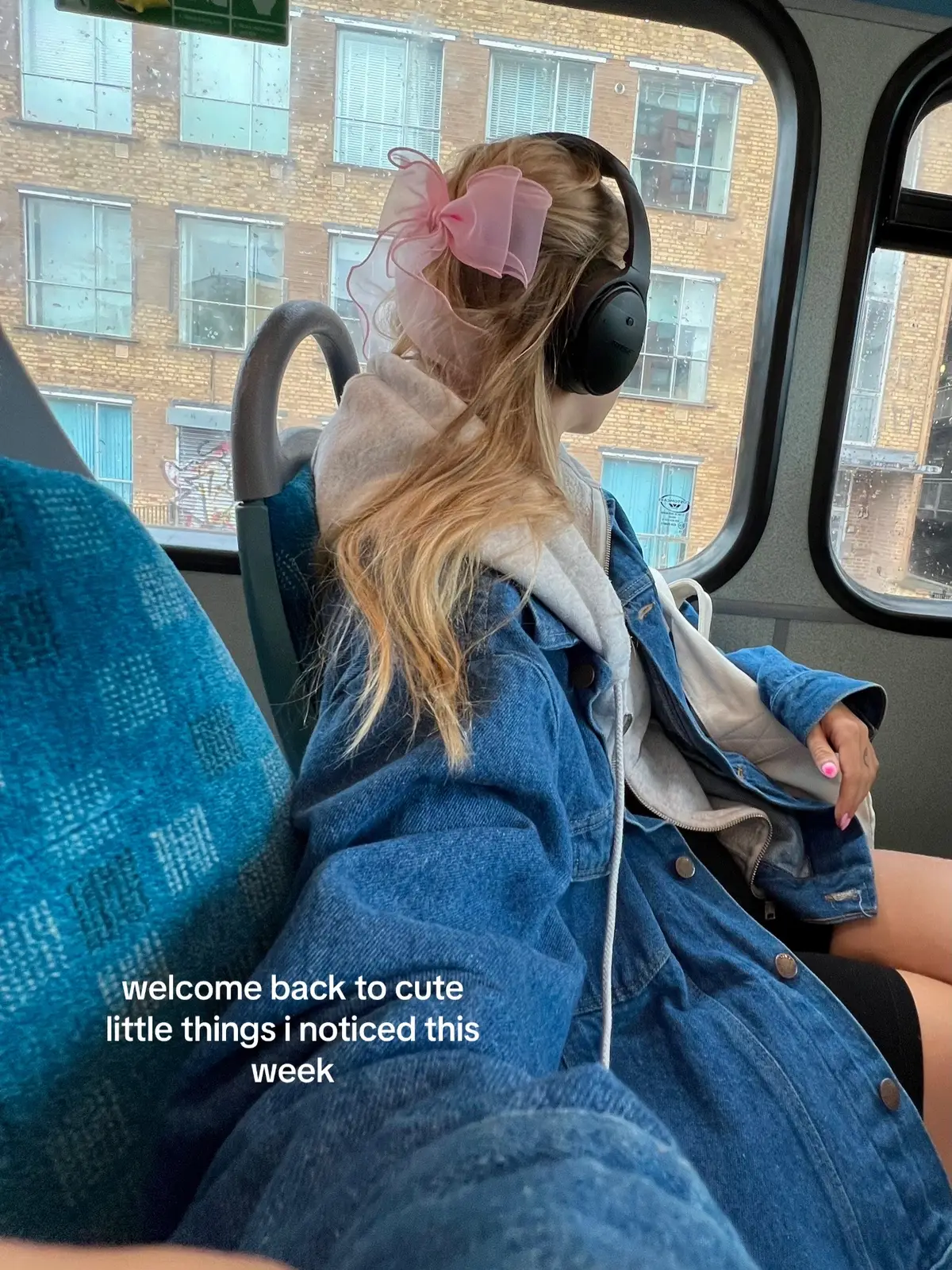 A person with blonde hair sits on a public transport, looking out of a window. They wear a denim jacket, and a pink bow sits on their head. The window shows an urban setting with buildings.