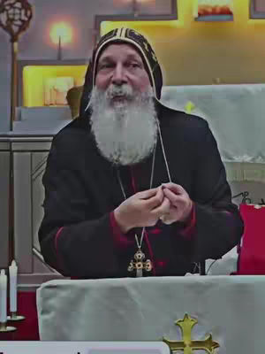 An older man with a white beard and a black religious robe is the main focus. He is wearing a traditional head covering and a cross necklace. He is facing the camera with hands clasped near his chest. The background features a religious setting with candles and a golden cross visible.