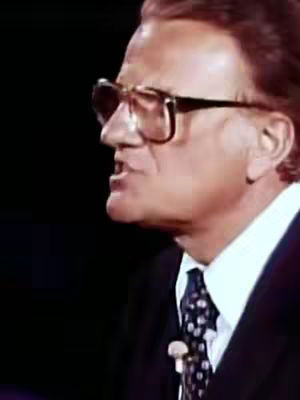 The image features a medium shot of Billy Graham, seen in profile from the side. He's wearing eyeglasses, a dark suit, a white collared shirt, and a patterned tie secured with a small microphone clip near his neck. His mouth is open, suggesting he is speaking. The background is a deep black.