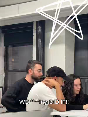 Multi-person scene in an office setting. Three figures are present: a man with glasses and beard, a person wearing a white shirt and a black cap, and a woman. The person in the white shirt is positioned centrally, with their head down. A white star-shaped design is overhead. The background includes a window and a wall with office-like features.