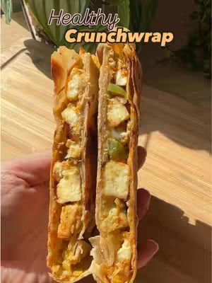 The image shows a close-up of a crunchy wrap cut in half, revealing the filling. The wrap is golden brown and flaky. The filling includes white paneer cubes, green bell peppers, and other vegetables. The background is a wooden cutting board, and a hand is holding the wrap. The top left is labeled "Healthy Crunchwrap.