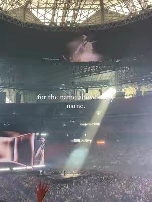 An eye-level shot captures a large indoor event, likely a conference or concert, with a stage setup at the bottom and a massive screen displaying a performer. A bright beam of light shines down, illuminating the scene from above, while a crowd reaches towards the stage. The image is dominated by dark tones with vibrant stage lighting.
