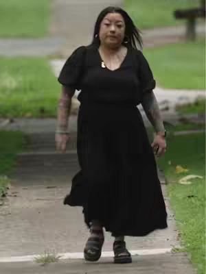 A woman wearing a black dress and sandals walks down a sidewalk. The background features green grass and trees.