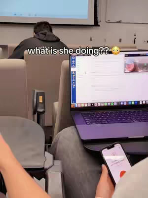 The scene is set in a lecture hall. A person in the back is turned away from the camera, and a laptop is open, showing a screen displaying what appears to be an online video. A cell phone is held up in front of the camera, suggesting a live stream or recording in progress. Text overlay displays the question: what is she doing??