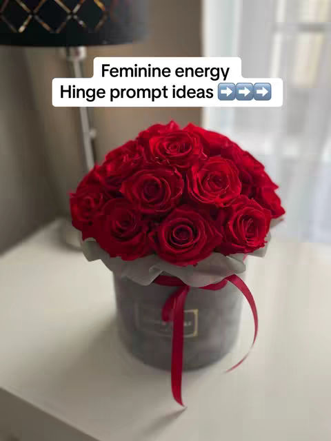 A close-up shot of a bouquet of red roses in a gray, cylindrical box with a red ribbon, in front of a blurred background that may be a room. A text overlay reads 'Feminine energy hinge prompt ideas' above the flowers.