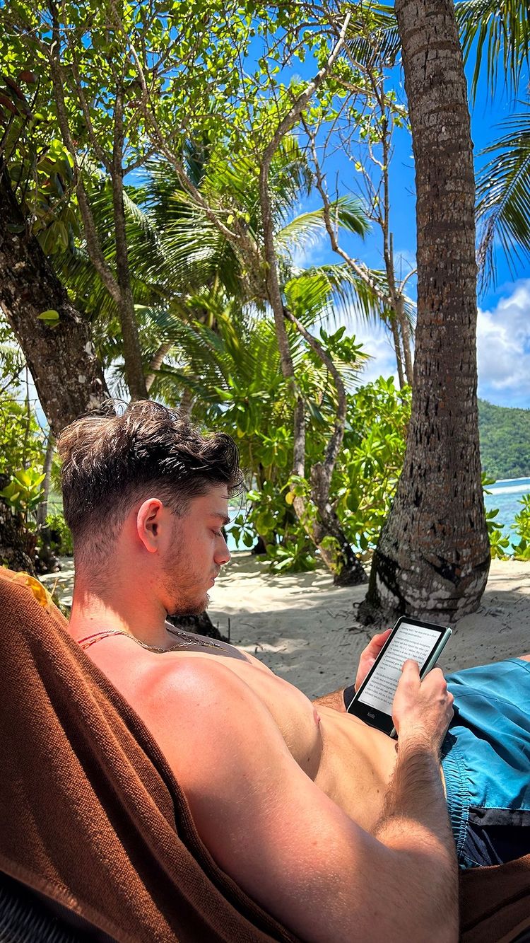 A man is relaxing on a beach, basking in sunlight and reading an e-reader. Lush green trees and palm trees provide shade, while a bright blue sky and ocean are visible in the background, creating a relaxed, tropical ambiance.