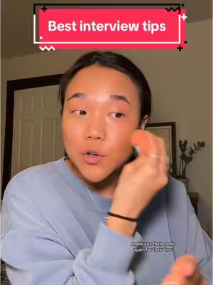 A woman is in the frame, mid-shot, with a softly lit complexion. She is wearing a powder blue top and has her hair styled back. She is holding in her right hand what appears to be a makeup sponge near her cheek and her left hand is closer to the camera. Above her head, a red bar overlay reads 'Best interview tips' in a clean, white font.