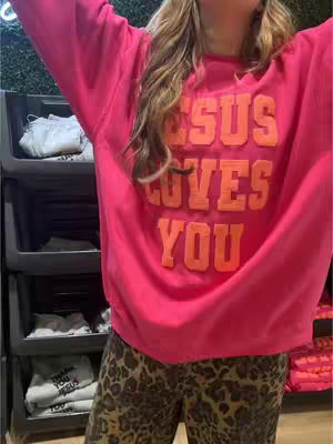 A woman wearing a bright pink sweater with the words "JESUS LOVES YOU" in orange block letters is the central focus. The sweater is paired with leopard print pants. The background includes a black retail shelving unit with items inside.