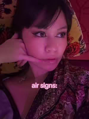 A young woman with dark hair poses with her hand near her face in a 'call me' gesture, set against a backdrop of a flowered blanket, illuminated by a pinkish-red light and a text overlay with the words 'air signs:'.