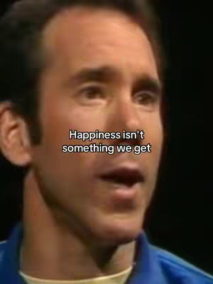 A medium shot captures a man with short brown hair, appearing to be in his late middle age. He is wearing a blue collared shirt. The background is dark and out of focus. Overlayed on the man's image is white text within a black rectangle that states, 'Happiness isn't something we get'.