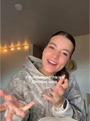 A smiling woman with fair skin and dark hair is in front of a camouflaged hoodie. Text is overlaid, and the background features a neutral tones setting with a light fixture visible.