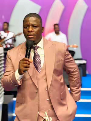 A man in a pink suit holds a microphone and speaks in what appears to be a church. Two people are blurred in the background, one playing a guitar. The background is purple with white pillar-like shapes. The lighting is bright with a warm tone.