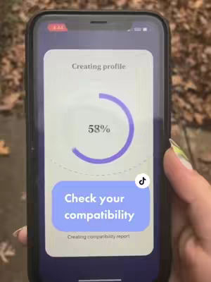 The image shows a close-up view of a smartphone screen held by a person, likely a female, with a manicured hand. The phone displays an app interface with a progress bar indicating a profile is being created at 58%. Overlaid on the screen are the words 'Check your compatibility' inside a blue rectangular button and a white TikTok logo. The background is blurred, featuring a mix of browns and greens suggestive of an outdoor setting.