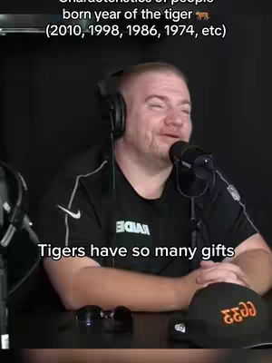 A man with a shaved head wearing a black t-shirt and headphones is speaking into a microphone. Text overlays are visible at the top and bottom. The background is a dark, undefined space. A black hat with orange lettering and a pair of sunglasses are on the desk.