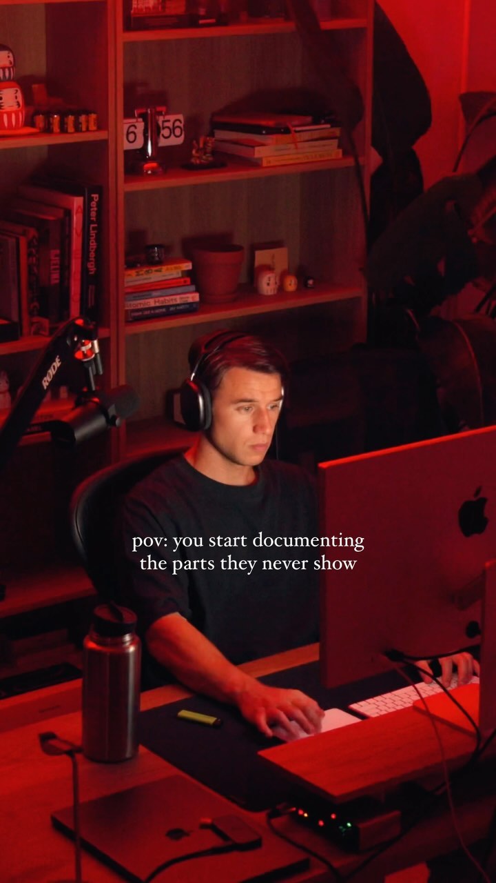 A man is seated in a dimly lit room with a desk, shelves, and a computer with the text overlayed.