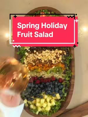 An overhead shot displays a fruit salad artfully arranged in a wooden, oval-shaped platter. The salad is composed of various layers of colorful fruits and toppings. A red rectangular text overlay reads "Spring Holiday Fruit Salad". A hand with a clear container holding liquid is slightly visible at the left, suggesting someone is either making the salad or adding a dressing.