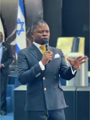 A medium shot of a man in a dark suit addressing an audience. He holds a microphone, with his right arm gesturing outward. Behind him, a flag with a Star of David is faintly visible. The lighting is bright, and the background suggests an indoor event.