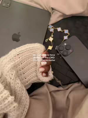 An overhead shot features a hand in a cream-colored sweater, holding an iPhone. Adjacent lies a dark grey iPad with the Apple logo. A gold bracelet with blue flower-shaped charms is positioned on the right. The composition rests on a bed covered in beige sheets. Text overlayed in white reads: 'don't attempt to force perfection. the LORD LOVES YOU no matter what.'
