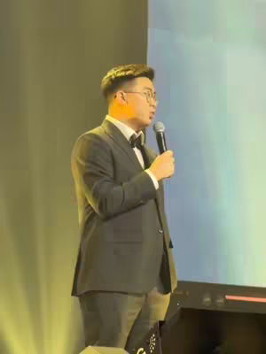 The cover image shows a man in a suit speaking into a microphone on a stage. The background is a blurred light-colored gradient. The man is positioned slightly off-center, with a focused expression.