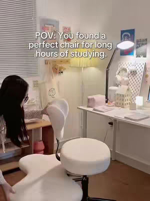 A young woman is shown partially from behind in a well-lit room, with a focus on a cream-colored ergonomic chair that is the apparent subject of the video. The chair has a unique shape with a rounded seat and back support. Sunlight streams in from a window, illuminating the room and highlighting the desk and its contents. The scene has a warm and inviting feel, characteristic of a home office or study space.