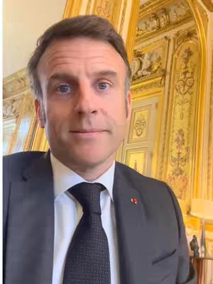 A medium, slightly upward-angle shot of a man in a suit. He is looking directly at the camera with a slight smile. The background shows an ornate, gold-colored wall.