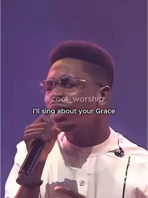 A man with short dark hair, wearing glasses and a white shirt, is holding a microphone and singing. The background is blurred purple.