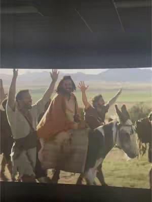 The image captures a scene from a movie featuring three men waving with joy, with one riding a donkey, set against a backdrop of a field and mountains, likely a biblical scene. A dark bar is present on top of the image