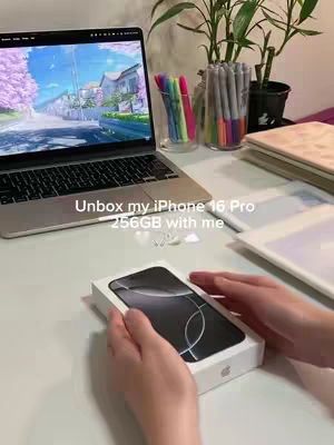 A person's hands are holding the white box of an iPhone, other objects are sitting on a surface: a laptop displaying an animated background, a pot with a plant on it, a glass with markers, and a notebook.