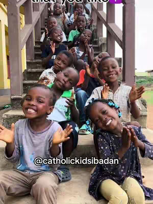 A group of diverse children, smiling, are sitting on a set of outdoor stairs. The top of the stairs are out of frame. One child is at the front, another slightly behind, with other children appearing behind them. They are all smiling up at the camera. The stairs are made of brown wood, and the background includes green plants. Text in the image says '@catholickidsibadan'. The lighting appears to be an overcast day. The overall composition is warm and inviting, highlighting the children's joyful expressions.