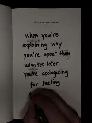 An open notebook displays handwritten text on a white page against a dark backdrop. The text is centered and appears to be a self-reflective quote about apologizing for one's feelings. A hand with a pen is visible at the bottom, as if in the process of writing.