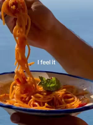 A close-up view showcases a hand holding up a portion of spaghetti with a vibrant red sauce, with some strands of spaghetti dangling down into a plate filled with more spaghetti. The background is a blurred sky and sea.