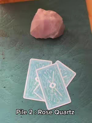 An overhead shot features a pile of three tarot cards in the lower center and a light pink rose quartz crystal in the upper center of the frame. The cards are light turquoise, and the crystal is also a pale color. The composition is completed with a matching background, completing a soft color palette.