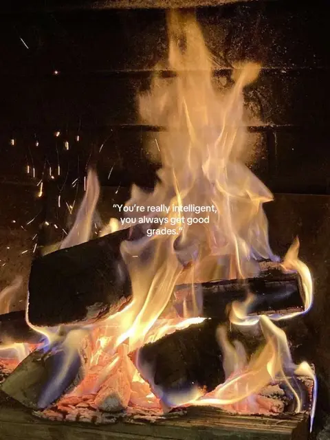 A close-up shot captures a roaring fire within a fireplace. Flames in shades of orange, yellow, and white dance above burning logs, some showing visible wood grain. Embers and sparks appear within and around the flames, suggesting intense heat. A text overlay reads, "You're really intelligent, you always get good grades." positioned against the dark background of the fireplace interior.