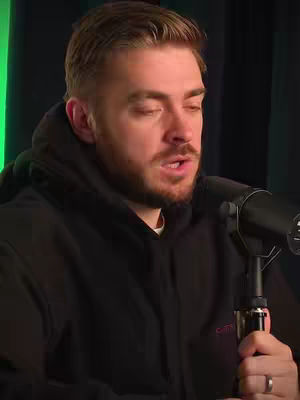 A medium shot shows a man, head and shoulders, in a black hooded sweatshirt. He is speaking into a microphone. The background is a dark, indistinct space that has a bright strip of green light on the left.